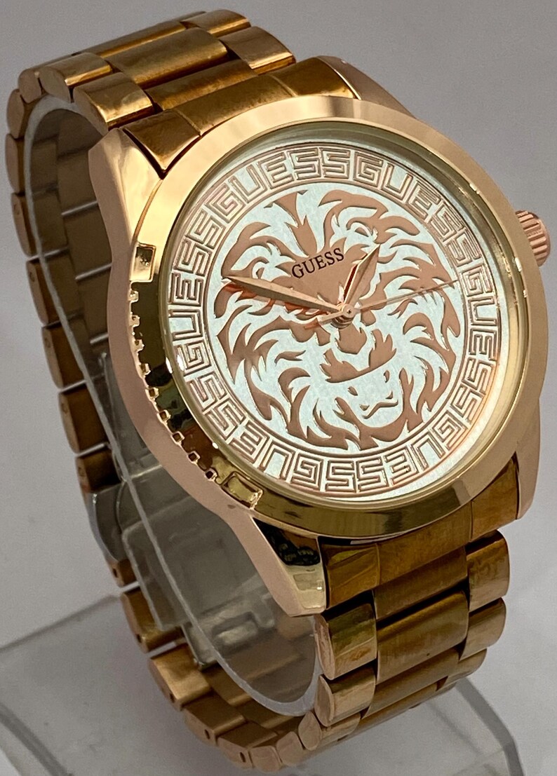 May include: Gold-toned Guess watch with a round face featuring a lion design. The watch has a gold band and a textured bezel. The watch face has the word "GUESS" and a lion graphic.