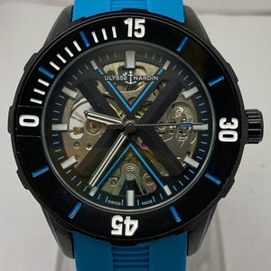 May include: A Ulysse Nardin skeleton watch with a black case and a bright blue rubber strap. The dial features a prominent black X-shaped bridge, white hour markers, and a rotating black bezel with white numerals 15, 30, and 45.