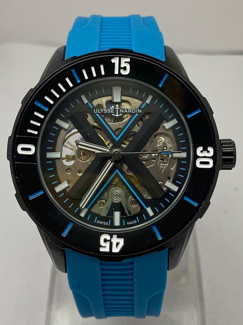 May include: A Ulysse Nardin skeleton watch with a black case and a bright blue rubber strap. The dial features a prominent black X-shaped bridge, white hour markers, and a rotating black bezel with white numerals 15, 30, and 45.
