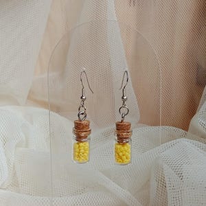 Op de afbeelding: Een paar unieke oorbellen met kleine glazen flesjes gevuld met gele kralen. Elk flesje is afgesloten met een kurk en hangt aan een zilverkleurige haak. De oorbellen worden gepresenteerd op een transparante standaard.