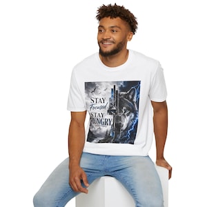 Op de afbeelding: Een wit t-shirt met korte mouwen en een grafische print van een wolvenkop, verdeeld tussen een berglandschap en een stormachtige lucht met blauwe bliksem. De tekst "STAY FOCUSED STAY HUNGRY" staat in een donker lettertype aan de linkerkant.