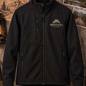 Könnte beinhalten: Eine schwarze Softshelljacke mit durchgehendem Frontreißverschluss, hohem Kragen und zwei unteren Reißverschlusstaschen. Auf der rechten Seite befindet sich eine vertikale Brusttasche mit Reißverschluss. Der Text MONTANA FLOW mit Berggrafik ist beige aufgedruckt.