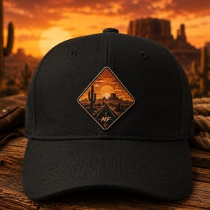 May include: A black baseball cap featuring a diamond-shaped patch on the front. The patch displays a desert landscape at sunset with a road, cacti, and rock formations, with the letters MF printed in a stylized font at the bottom of the design.