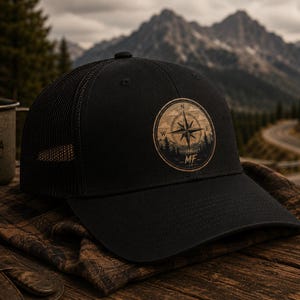 Pode incluir: Um boné trucker preto com um emblema circular na frente. O emblema apresenta um design de rosa dos ventos com as letras MF na parte inferior, rodeado por uma silhueta de floresta. O boné tem um painel frontal sólido e rede atrás.