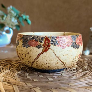 Puede incluir: Un cuenco de cerámica beige con un estampado floral en rojo, azul y negro. El cuenco tiene grietas visibles reparadas con laca dorada, mostrando el arte japonés del Kintsugi. El cuenco está sobre una esterilla tejida.