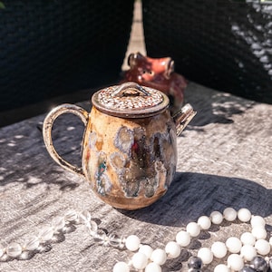 May include: A small ceramic teapot with a speckled tan and brown glaze featuring a textured lid with a small handle. The body of the teapot has a mottled finish with hints of blue and dark brown tones, a curved side handle, and a short spout.