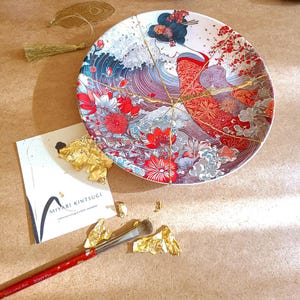 May include: A round ceramic plate featuring a Kintsugi repair with gold lacquer lines. The plate displays a graphic illustration of a person in a red patterned kimono amidst stylized blue waves and red flowers. A brush and gold leaf are nearby.