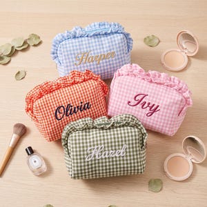 Puede incluir: Cuatro neceseres rectangulares con bordes de volantes y estampados de cuadros vichy en azul, naranja, rosa y verde. Cada neceser presenta nombres bordados: Harper en dorado, Olivia en azul oscuro, Ivy en magenta y Hazel en blanco.