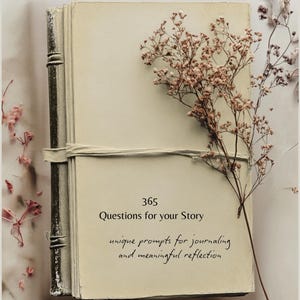 May include: A cream-colored journal with a dark, textured spine and a fabric tie closure. The cover features the text 365 Questions for your Story and unique prompts for journaling and meaningful reflection, accented by a sprig of dried flowers.