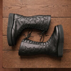 May include: A pair of black mid-calf boots featuring a thick platform sole and a lace-up front closure. The outer side of each boot is decorated with three stitched star patterns. The boots are constructed with smooth black material and black laces.