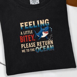 May include: Black t-shirt with the text "Feeling a little bitey, please return me to the ocean" and a cartoon shark graphic. The shirt is made of a soft material and is perfect for casual wear.