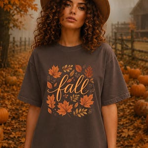May include: A dark grey short-sleeved t-shirt featuring a central graphic of orange and brown autumn leaves surrounding the word fall written in a cursive script font. The shirt has a crew neckline and a relaxed fit, showcasing a seasonal design.