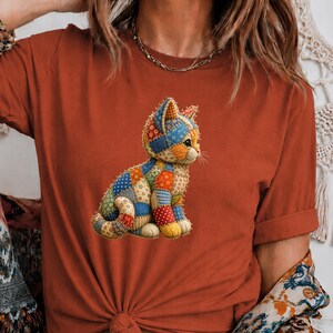 May include: A rust-colored short-sleeve t-shirt featuring a central graphic of a sitting cat composed of various colorful patchwork fabric patterns including blue, orange, yellow, and white dots, stripes, and floral motifs on the feline figure.