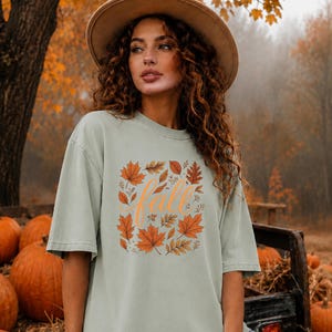 May include: A sage green short-sleeve t-shirt featuring a central graphic of orange and brown autumn leaves surrounding the word fall written in a cursive orange script. The shirt has a crew neckline and a relaxed, loose-fitting silhouette.