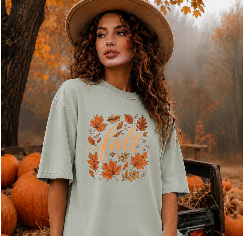 May include: A sage green short-sleeve t-shirt featuring a central graphic of orange and brown autumn leaves surrounding the word fall written in a cursive orange script. The shirt has a crew neckline and a relaxed, loose-fitting silhouette.