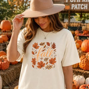 May include: A cream-coloured short-sleeve t-shirt featuring a central graphic of orange and brown autumn leaves surrounding the word fall in cursive script. The shirt is paired with a wide-brimmed tan felt hat and a delicate silver chain necklace.