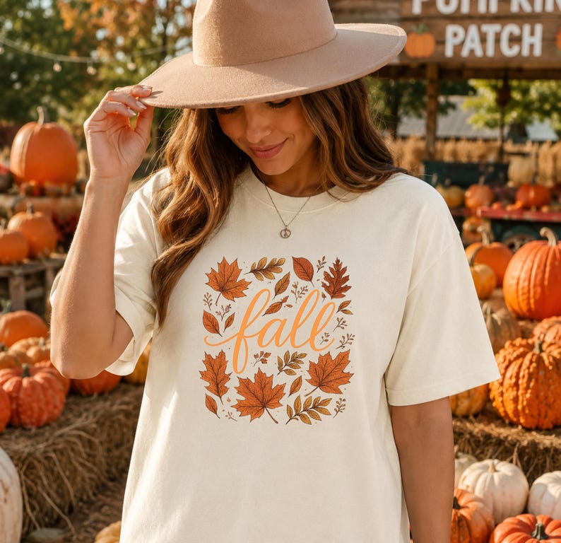 May include: A cream-coloured short-sleeve t-shirt featuring a central graphic of orange and brown autumn leaves surrounding the word fall in cursive script. The shirt is paired with a wide-brimmed tan felt hat and a delicate silver chain necklace.