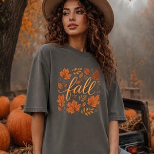 May include: A charcoal grey short-sleeve t-shirt featuring a central graphic of orange and yellow autumn leaves surrounding the word fall written in a cursive script. The shirt has a crew neckline and a relaxed fit with a faded fabric texture.