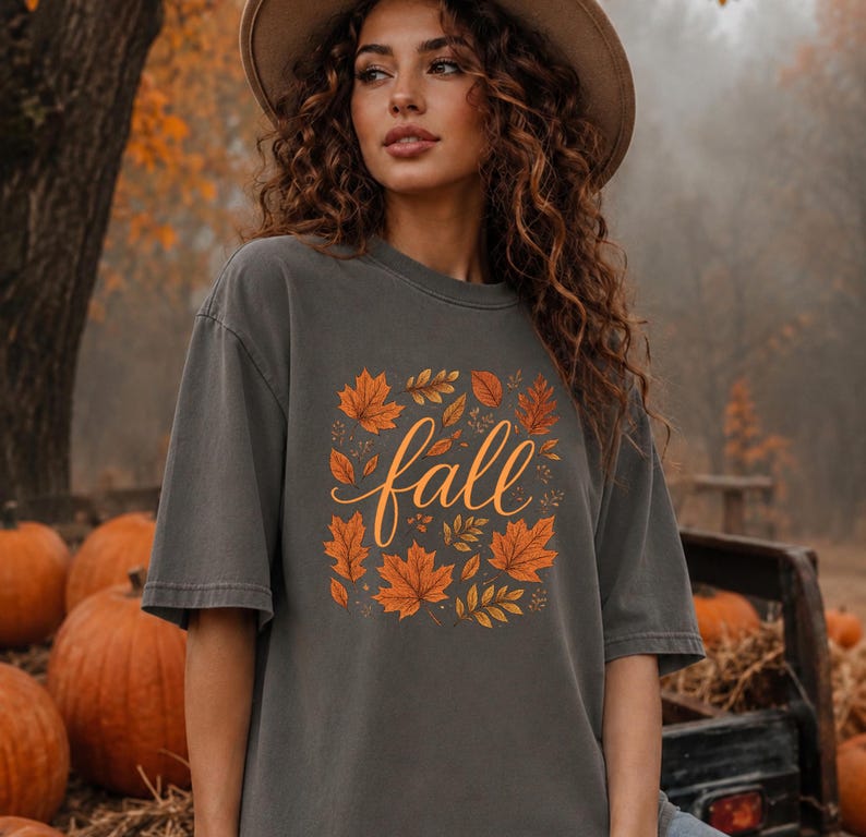 May include: A charcoal grey short-sleeve t-shirt featuring a central graphic of orange and yellow autumn leaves surrounding the word fall written in a cursive script. The shirt has a crew neckline and a relaxed fit with a faded fabric texture.