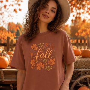 May include: A rust-coloured short-sleeve t-shirt featuring a central graphic of various autumn leaves in shades of orange, yellow, and brown surrounding the word fall written in a cursive, light-coloured script font across the chest area.