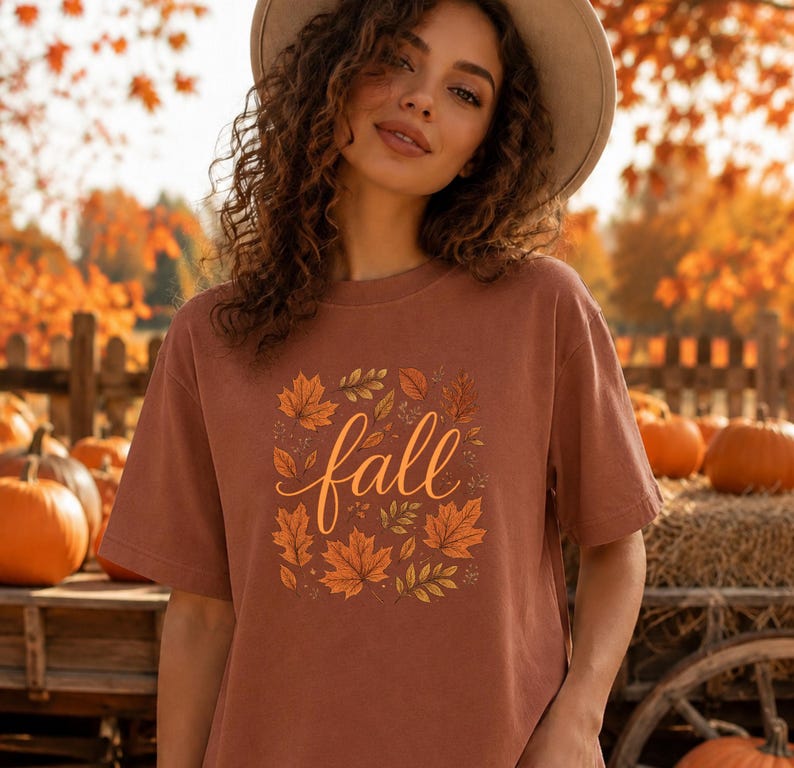 May include: A rust-coloured short-sleeve t-shirt featuring a central graphic of various autumn leaves in shades of orange, yellow, and brown surrounding the word fall written in a cursive, light-coloured script font across the chest area.