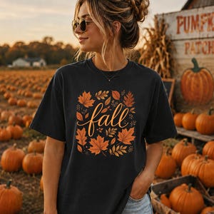 May include: A black short-sleeved t-shirt featuring a central graphic of orange and brown autumn leaves surrounding the word fall written in a cursive script. The shirt has a crew neckline and a relaxed fit, showcasing a seasonal design.