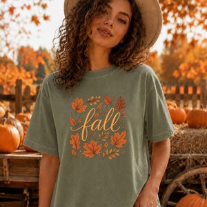 May include: A sage green short-sleeve t-shirt featuring a central graphic of the word fall in a cursive orange font, surrounded by a collection of various orange and brown autumn leaves arranged in a square pattern on the front of the garment.
