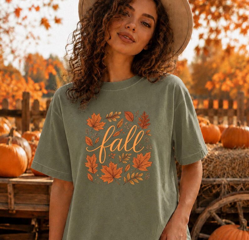 May include: A sage green short-sleeve t-shirt featuring a central graphic of the word fall in a cursive orange font, surrounded by a collection of various orange and brown autumn leaves arranged in a square pattern on the front of the garment.