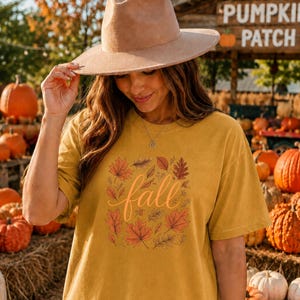 May include: A mustard yellow short-sleeve t-shirt featuring a central graphic of various autumn leaves in shades of orange, red, and brown surrounding the word fall in a cursive script font, paired with a wide-brimmed tan felt fedora hat.