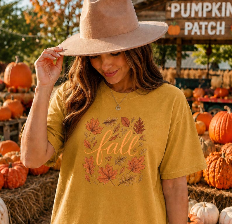 May include: A mustard yellow short-sleeve t-shirt featuring a central graphic of various autumn leaves in shades of orange, red, and brown surrounding the word fall in a cursive script font, paired with a wide-brimmed tan felt fedora hat.