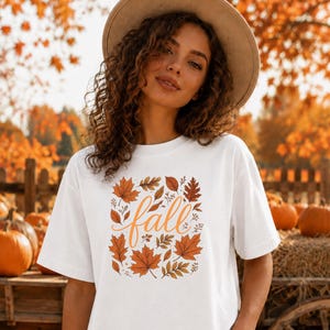 May include: A white short-sleeved t-shirt featuring a central graphic of the word fall in orange cursive script, surrounded by a variety of illustrated autumn leaves in shades of orange, brown, and gold, worn with a tan wide-brimmed hat.