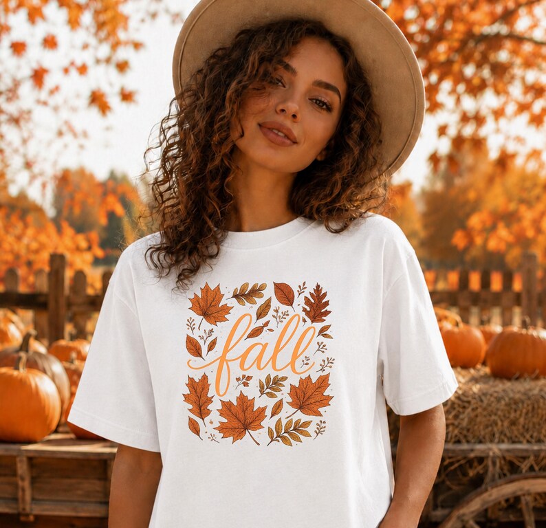 May include: A white short-sleeved t-shirt featuring a central graphic of the word fall in orange cursive script, surrounded by a variety of illustrated autumn leaves in shades of orange, brown, and gold, worn with a tan wide-brimmed hat.
