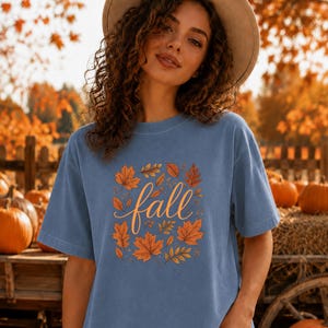 May include: A blue short-sleeved t-shirt featuring a central graphic of the word fall in a cursive script, surrounded by an arrangement of orange, yellow, and brown autumn leaves. The design is printed on the front of the cotton crewneck shirt.