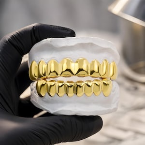 Może przedstawiać: Zestaw złotych grillów dentystycznych, prezentowany na białym odlewie dentystycznym. Grille są mocno wypolerowane i odbijają światło, prezentując jasne, metaliczne wykończenie. Zdjęcie zostało zrobione w studio, podkreślając kunszt akcesorium dentystycznego.