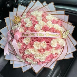 May include: A large bouquet of pink and white roses, wrapped in decorative paper. A pink ribbon with the text "Para Mi Ni&ntilde;a Hermosa!" is draped across the flowers. Gold-colored crown and butterfly accents add a touch of elegance.