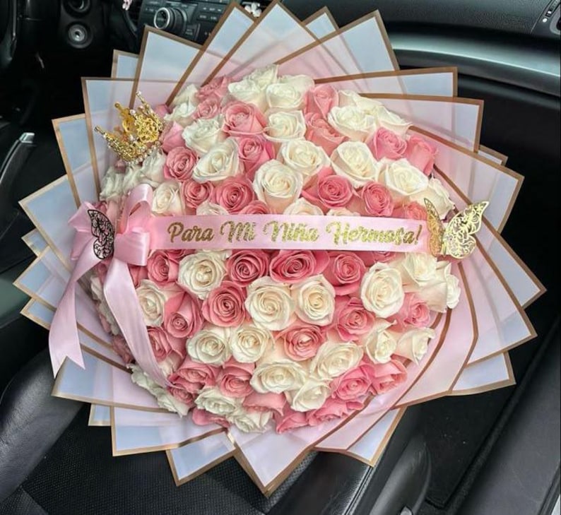 May include: A large bouquet of pink and white roses, wrapped in decorative paper. A pink ribbon with the text "Para Mi Ni&ntilde;a Hermosa!" is draped across the flowers. Gold-colored crown and butterfly accents add a touch of elegance.