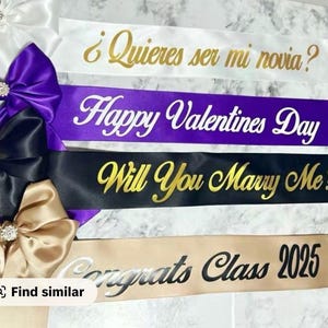 May include: Satin sashes in white, purple, black, and gold, with text in gold and white. The sashes have text that reads "&iquest;Quieres ser mi novia?", "Happy Valentines Day", "Will You Marry Me?", and "Congrats Class 2025". Each sash is adorned with a bow.