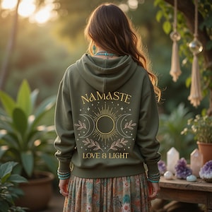 Puede incluir: Una sudadera con capucha de color verde oliva que presenta un gráfico en la espalda con el texto "NAMASTE" y "LOVE & LIGHT" rodeando un diseño central de sol y luna con detalles botánicos. La sudadera tiene mangas largas y capucha.