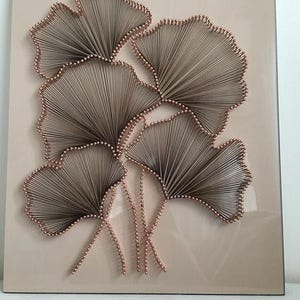 Puede incluir: Una pieza de arte de hilo hecha a mano con cinco hojas de ginkgo estilizadas. Las hojas están hechas con hilo marrón y alfileres color cobre, sobre un fondo rosa claro. Una decoración de pared única.
