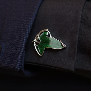 Puede incluir: Gemelo plateado con forma de hoja, con un diseño de esmalte verde vibrante. El gemelo presenta detalles de venas delicadas y se coloca sobre una chaqueta de traje azul oscuro, un accesorio clásico.