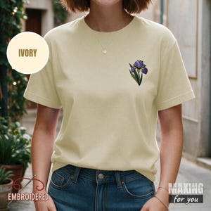 Könnte beinhalten: Ein elfenbeinfarbenes Kurzarm-T-Shirt mit einer kleinen gestickten violetten Irisblüte mit grünen Blättern auf der linken Brust. Das T-Shirt hat einen Rundhalsausschnitt und wird mit einer blauen Jeans kombiniert. Ein Etikett zeigt die Farbe Elfenbein.