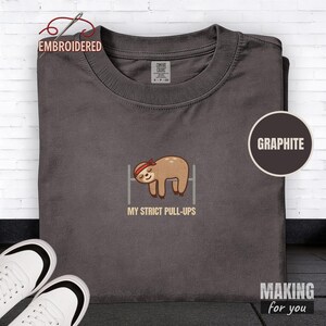 May include: A graphite grey crew neck t-shirt featuring an embroidered graphic of a brown sloth wearing a red headband while hanging from a horizontal bar. Below the sloth, the text reads MY STRICT PULL-UPS in a light tan, sans-serif font.