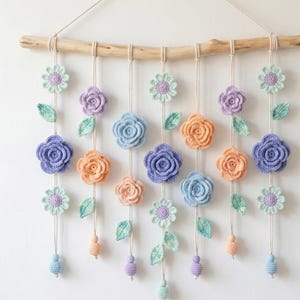 Puede incluir: Un colgante de pared floral hecho a mano con flores de ganchillo en tonos azules, naranjas, morados y verdes. Las flores están suspendidas de una rama de madera natural con cuerdas colgantes y cuentas decorativas, creando una pieza decorativa.