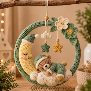 May include: A circular crochet wall hanging featuring a yellow crescent moon with a sleeping face and a brown teddy bear wearing a green cap and outfit, both resting on a white cloud. The hoop is decorated with green flowers and hanging stars.