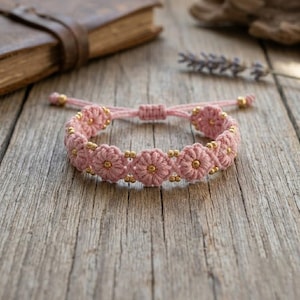 Puede incluir: Una pulsera de macramé rosa que presenta una serie de nudos interconectados con forma de flor. Cada centro de flor está acentuado con una pequeña cuenta de metal dorado, y hay cuentas doradas adicionales entre los motivos florales.