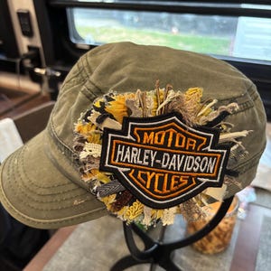 May include: Olive green military-style cap with a Harley-Davidson Cycles patch. The patch is black, orange, and white, with a decorative floral accent. The cap is displayed on a black stand.