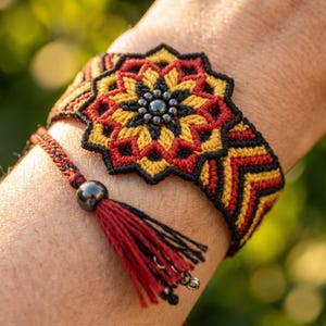 May include: A handmade bracelet featuring a central floral design in red, yellow, and black. The bracelet has a matching tassel with a black bead and red and black threads. The intricate macrame design is visible.