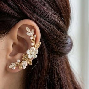 May include: A gold-toned ear cuff featuring a botanical design with textured leaves and white floral accents. The piece is embellished with small white pearls and clear crystals, following the curve of the ear from the lobe to the upper cartilage.