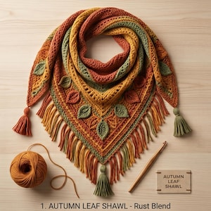 May include: A triangular crochet shawl in rust, mustard yellow, and sage green featuring leaf motifs and fringe. Includes a ball of matching yarn, a wooden crochet hook, and a rectangular tag labelled AUTUMN LEAF SHAWL. 1. AUTUMN LEAF SHAWL - Rust Blend.