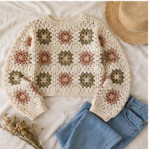 Könnte beinhalten: Cremefarbener Häkelpullover mit floralem Granny-Square-Muster in Grün- und Rosatönen. Der Pullover wird mit einer blauen Jeans und einem Strohhut kombiniert, was eine lässige, böhmische Ästhetik erzeugt. Trockenblumen sind ebenfalls vorhanden.